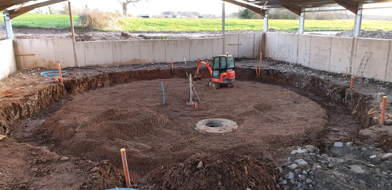 Circular excavation for a rotary parlour inside an open-sided farm building, with a mini digger at the edge